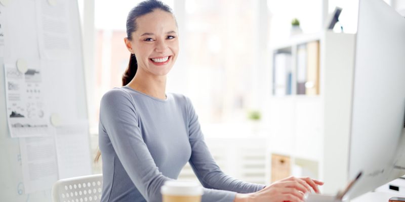 Young secretary looking at camera with toothy smile by her workplace while networking